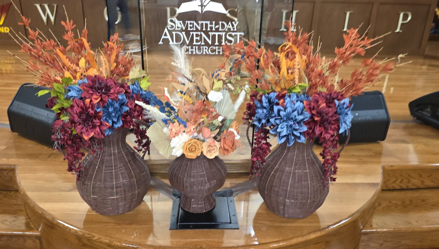 Three fall arrangements of rust, burgundy, and blue blooms in woven brown vases, set at the front of a church sanctuary
